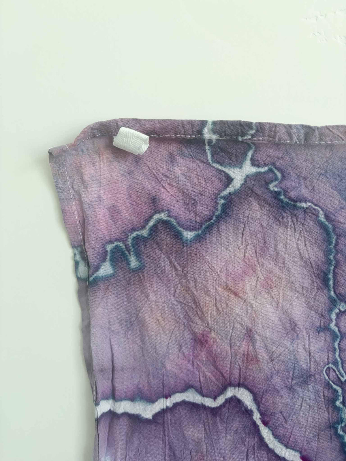 Corner of a violet and mauve ice dyed tea towel with a white twill hanging loop stitched into the hemmed edge, and white lines edged in slate blue.