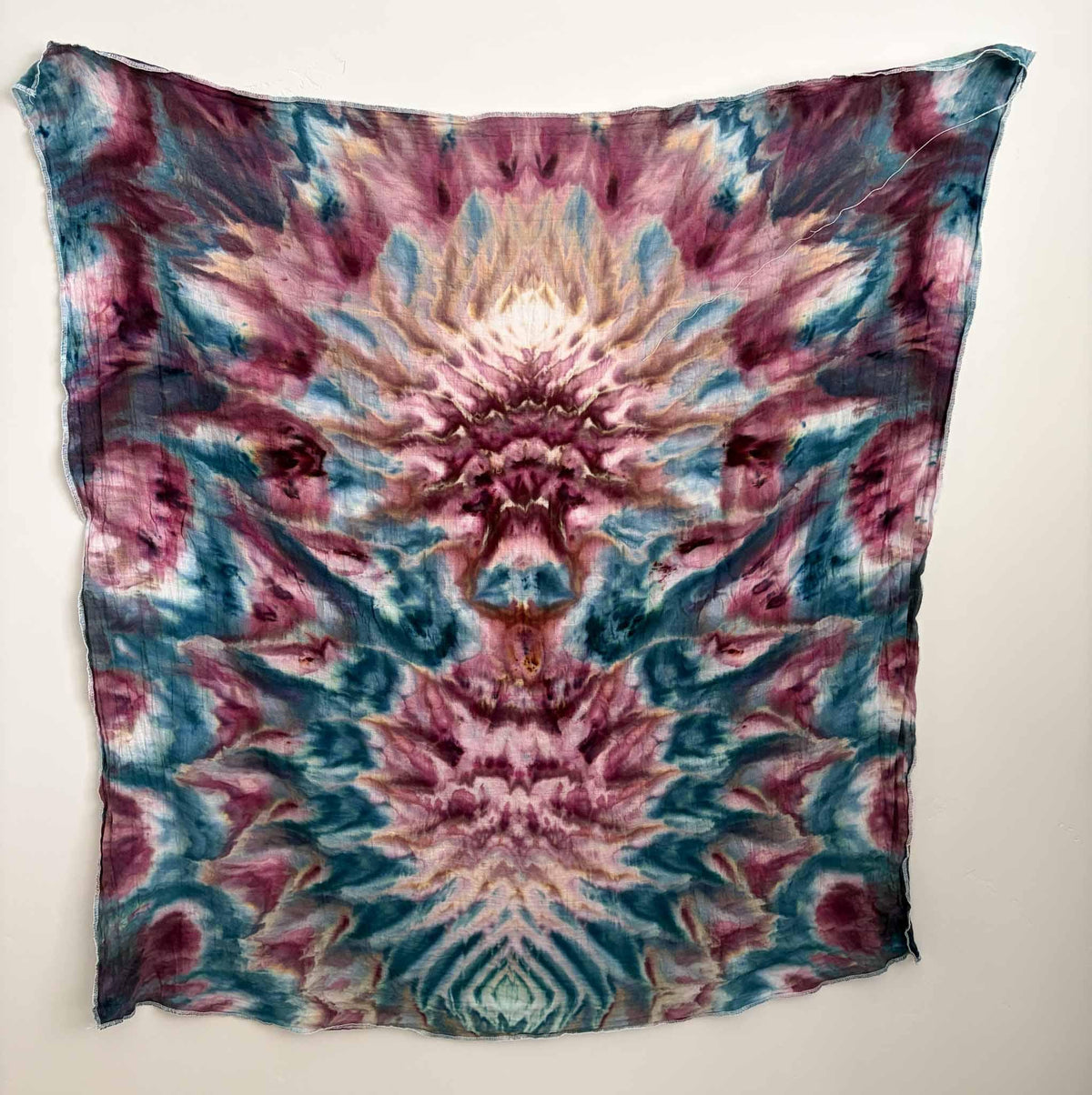 Square ice dyed bamboo gauze swaddle hanging on a white wall, showing a mirrored deity fold design in berry pink and teal blue with a cream and peach center.